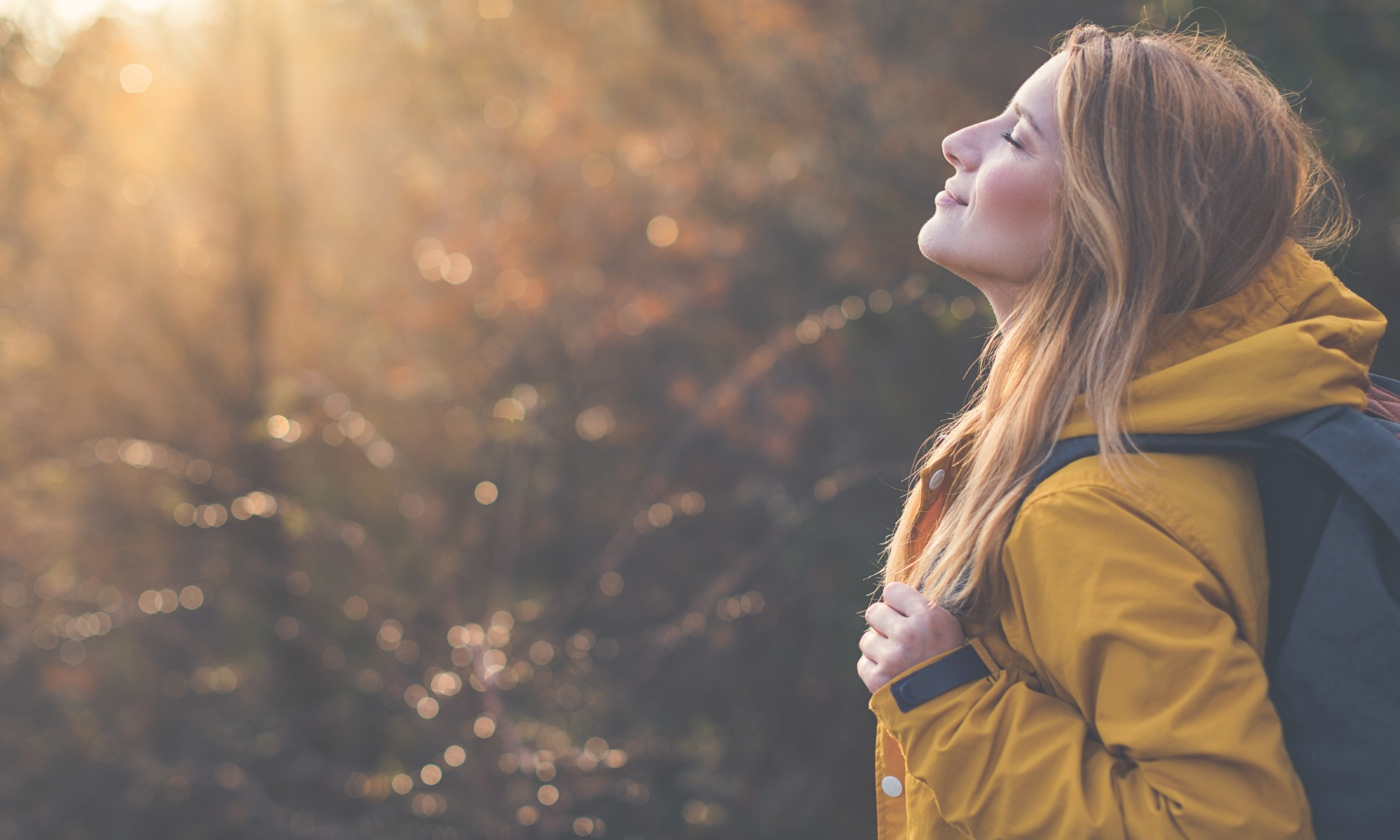 Nuori nainen keltaisessa takissa, reppu selässä, katsoo kohti aurinkoa loppukesän maisemassa